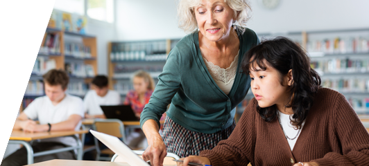 image of older woman in classroom helping student on laptop.